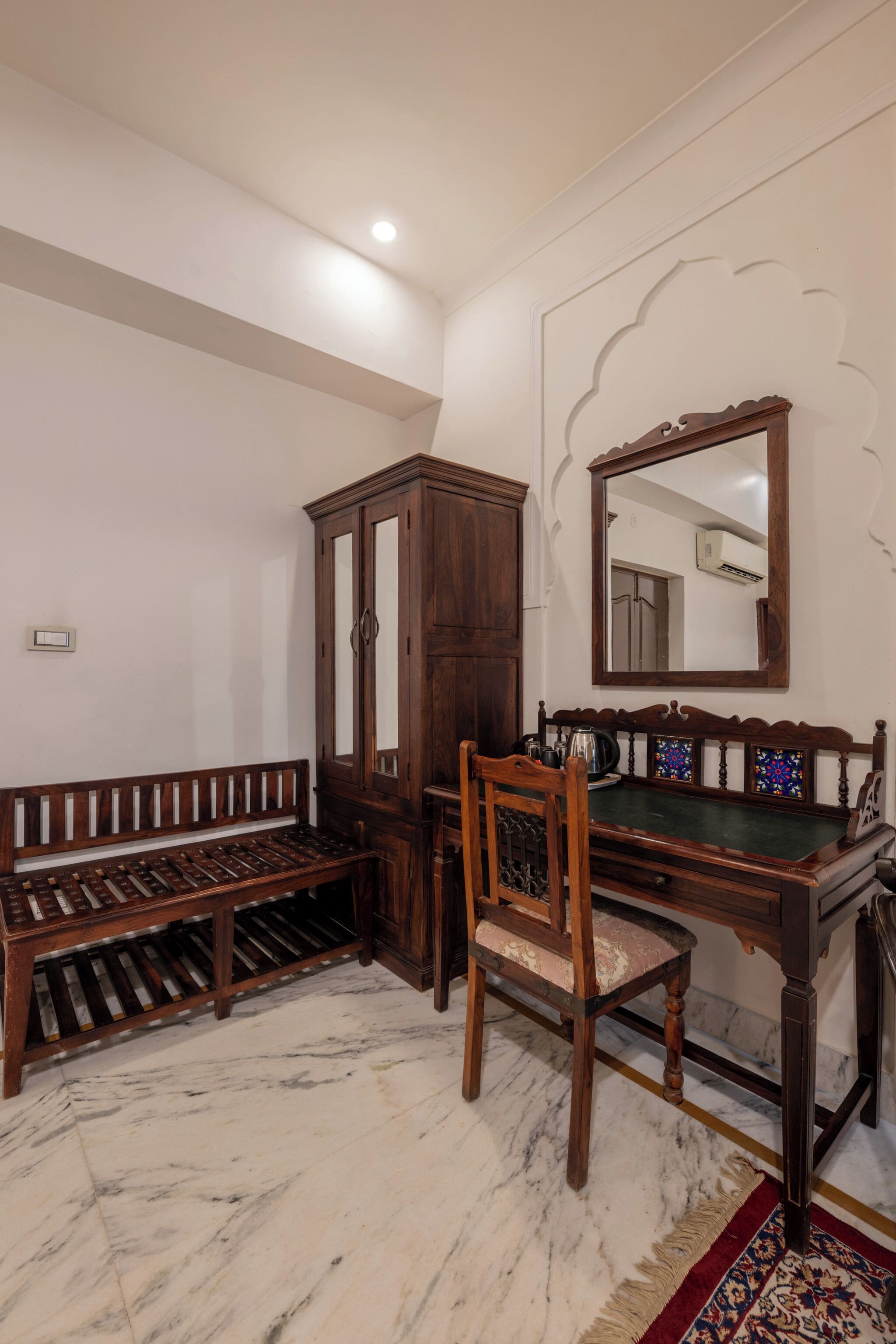 Um quarto com um banco de madeira, uma cadeira, um armário e um espelho na parede.