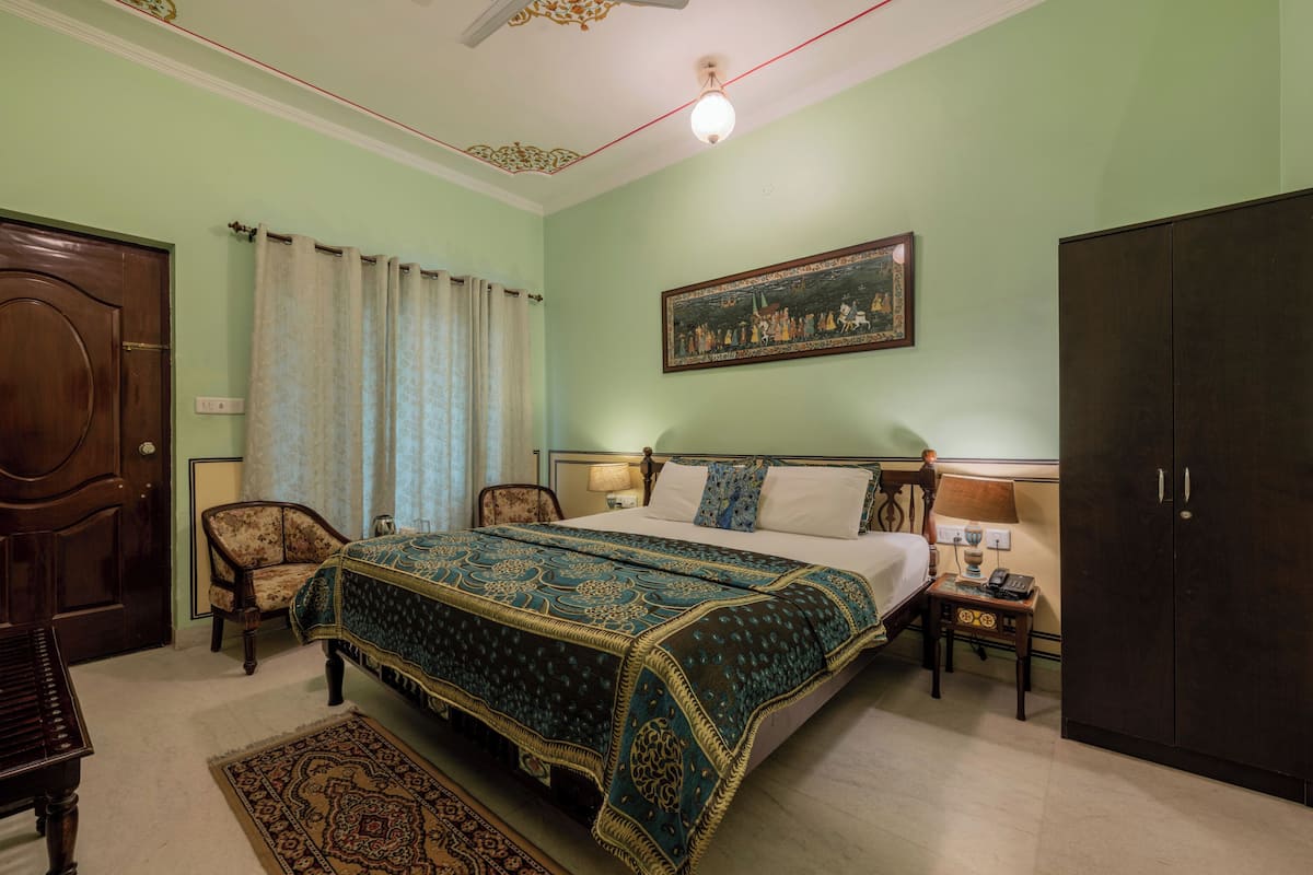 A bedroom with a large bed, a wooden wardrobe, a chair, a desk with a lamp, and a painting on the wall.