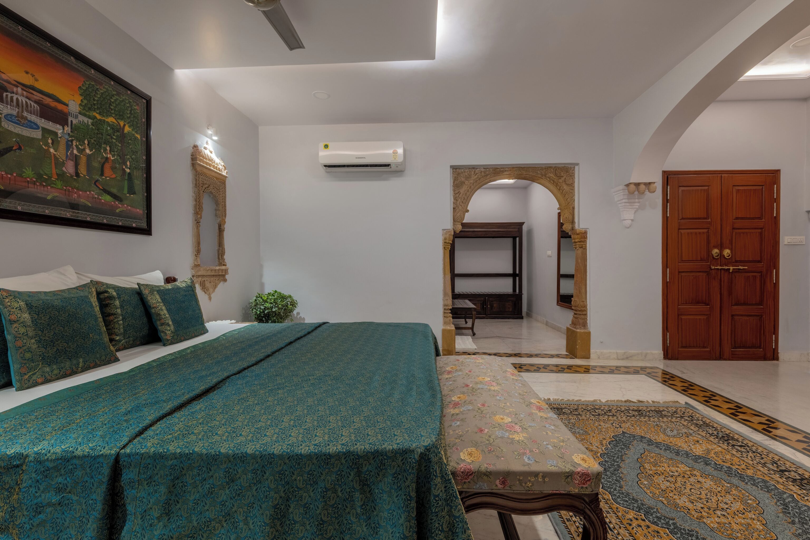 A bedroom with a bed, a painting, an air conditioner, a wooden door, and a patterned rug.