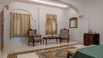 Una habitación con alfombra estampada, dos sillones, una mesa de centro de madera y una cama con cubierta verde.