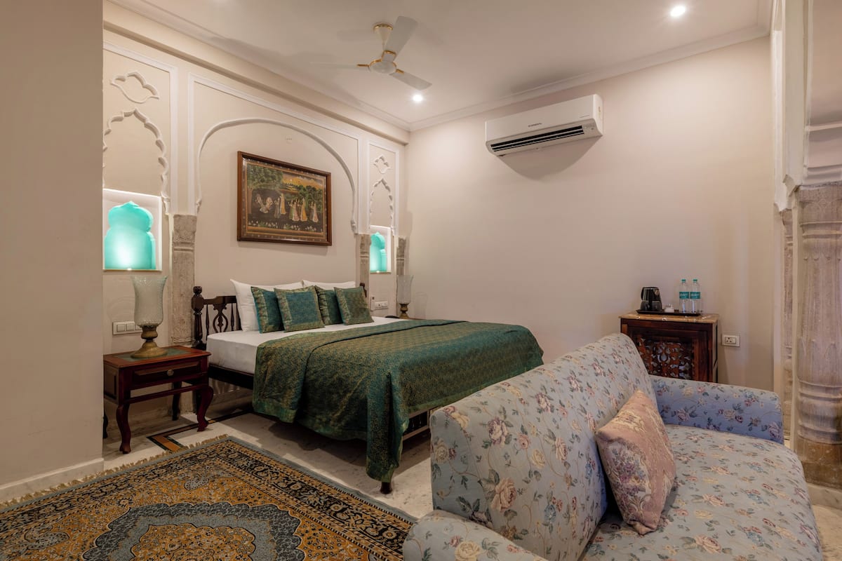 A bedroom with a bed, a sofa, a nightstand, a lamp, a painting, and an air conditioning unit.