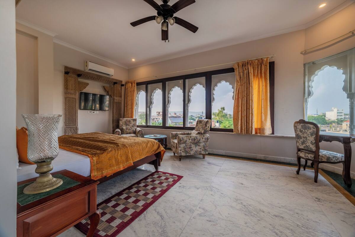 A bedroom with a bed, a desk, two chairs, a television, and a window with curtains.
