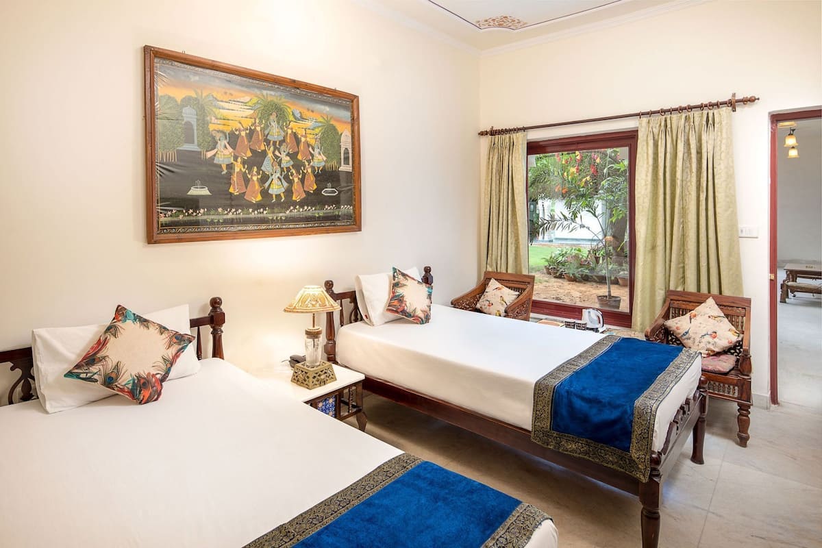 A hotel room with two beds, a painting, a window with curtains, and a view of greenery outside.