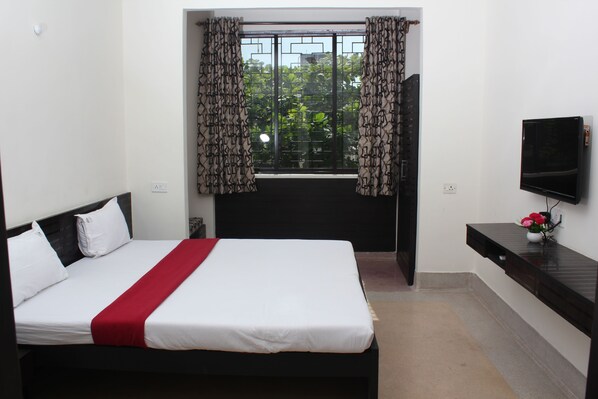 A hotel room with a bed, a TV, a window with curtains, and a small table with a vase.