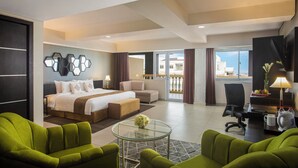A modern hotel room with a bed, green armchairs, a glass coffee table, a desk with a lamp, and a view of the outside. - Merapi Merbabu Hotel Yogyakarta Powered by Archipelago (Depok)