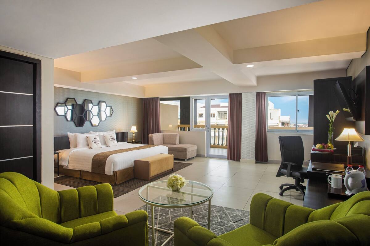 Une chambre d’hôtel moderne, équipée d’un lit, de fauteuils verts, d’une table basse en verre, d’un bureau avec une lampe et offrant une vue sur l’extérieur.