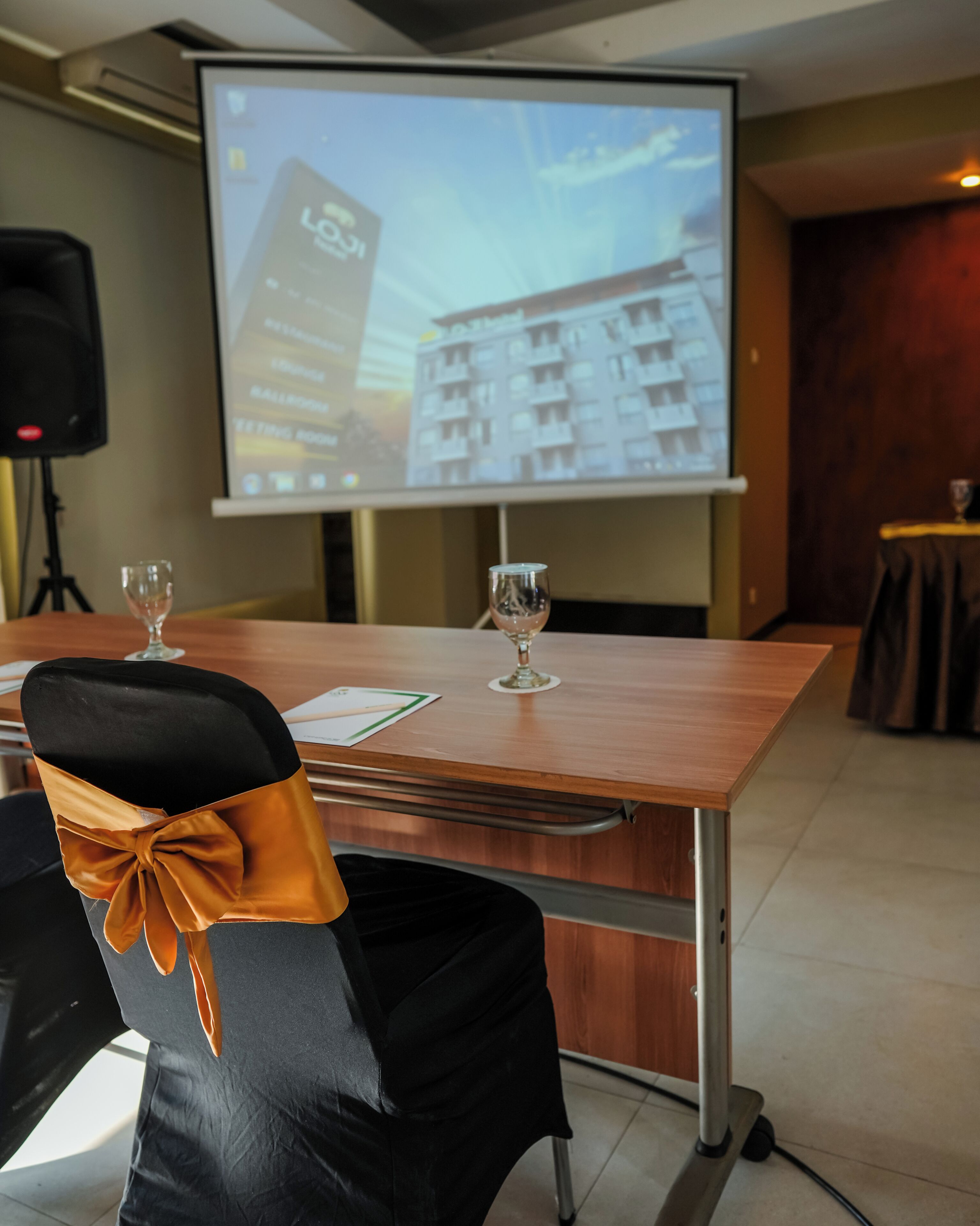 Un salón de conferencias con una pantalla de proyector mostrando una imagen de un edificio, dos copas de vino sobre la mesa y una silla decorada con un lazo.