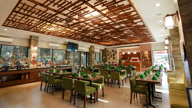 Un restaurant avec un plafond haut et des poutres en bois, des chaises recouvertes de tissu vert et un bar proposant une variété de plats.