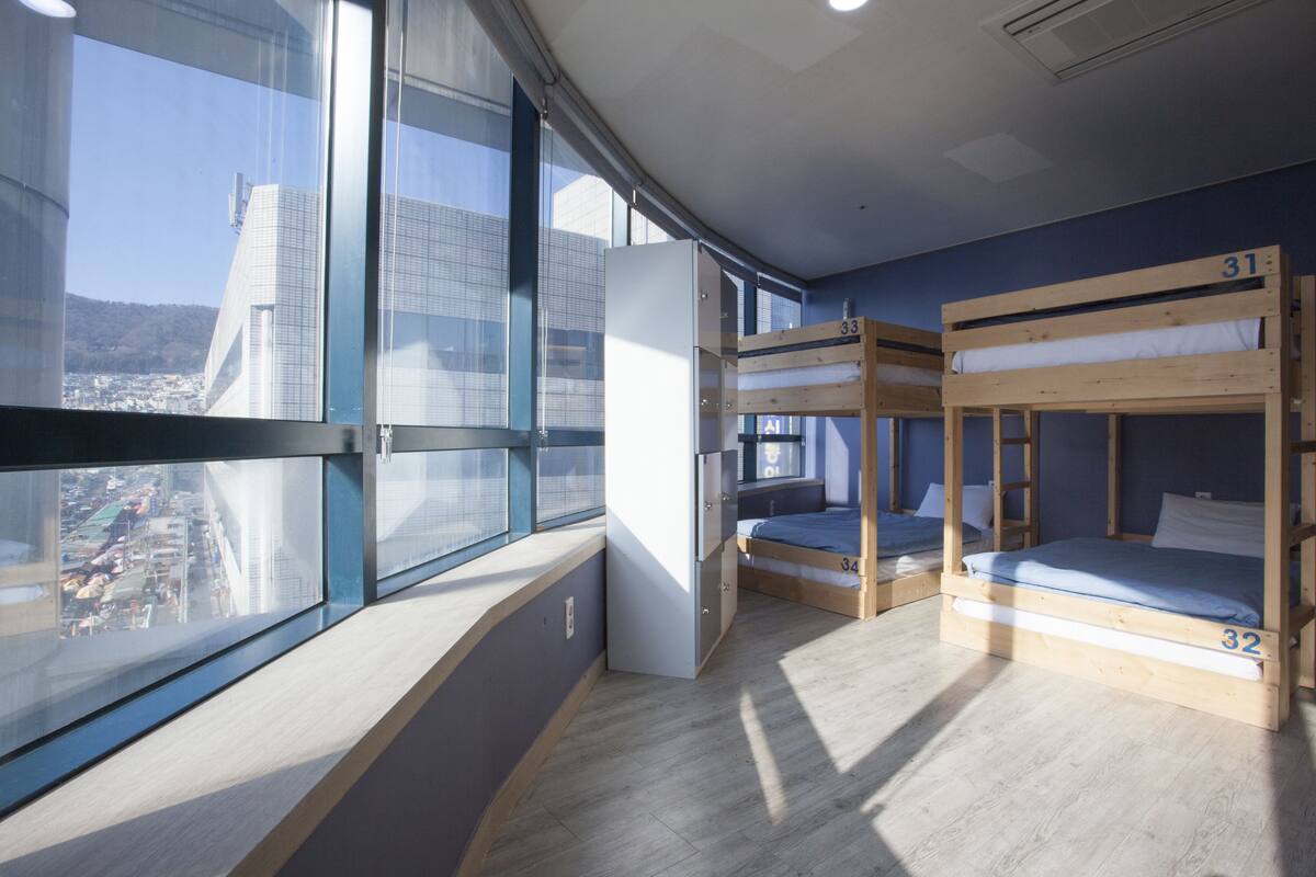 A dormitory room with bunk beds, a refrigerator, and a window with a city view.