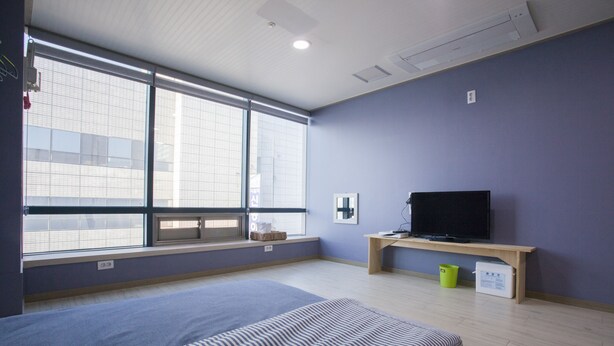 Habitación de hotel con un ventanal grande, una cama, un televisor sobre un mueble de madera y un escritorio pequeño.