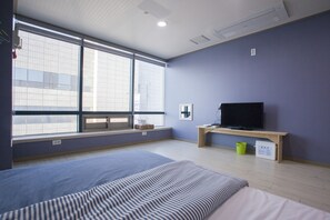 Habitación de hotel con un ventanal grande, una cama, un televisor sobre un mueble de madera y un escritorio pequeño.
