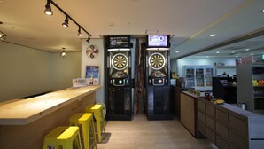 Una zona de bar con mostrador de madera, taburetes amarillos, dardos y una pantalla de televisión.