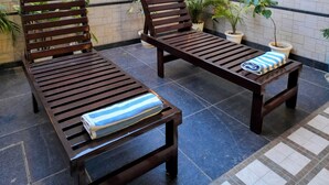 Un patio con dos sillones de madera y una mesita pequeña, cada uno con una toalla. El área está delimitada por un muro de ladrillo blanco con un panel decorativo de celosía.