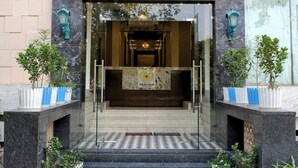 La entrada del hotel The Raj Vilas, con fachada de mármol, puertas de cristal y plantas en macetas.