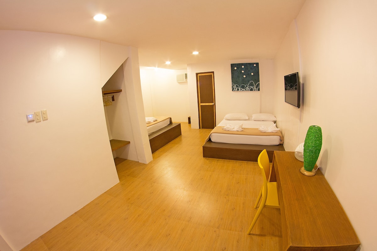 A hotel room with a wooden floor, a bed with white linens, a desk with a chair, a green vase, and a painting on the wall.