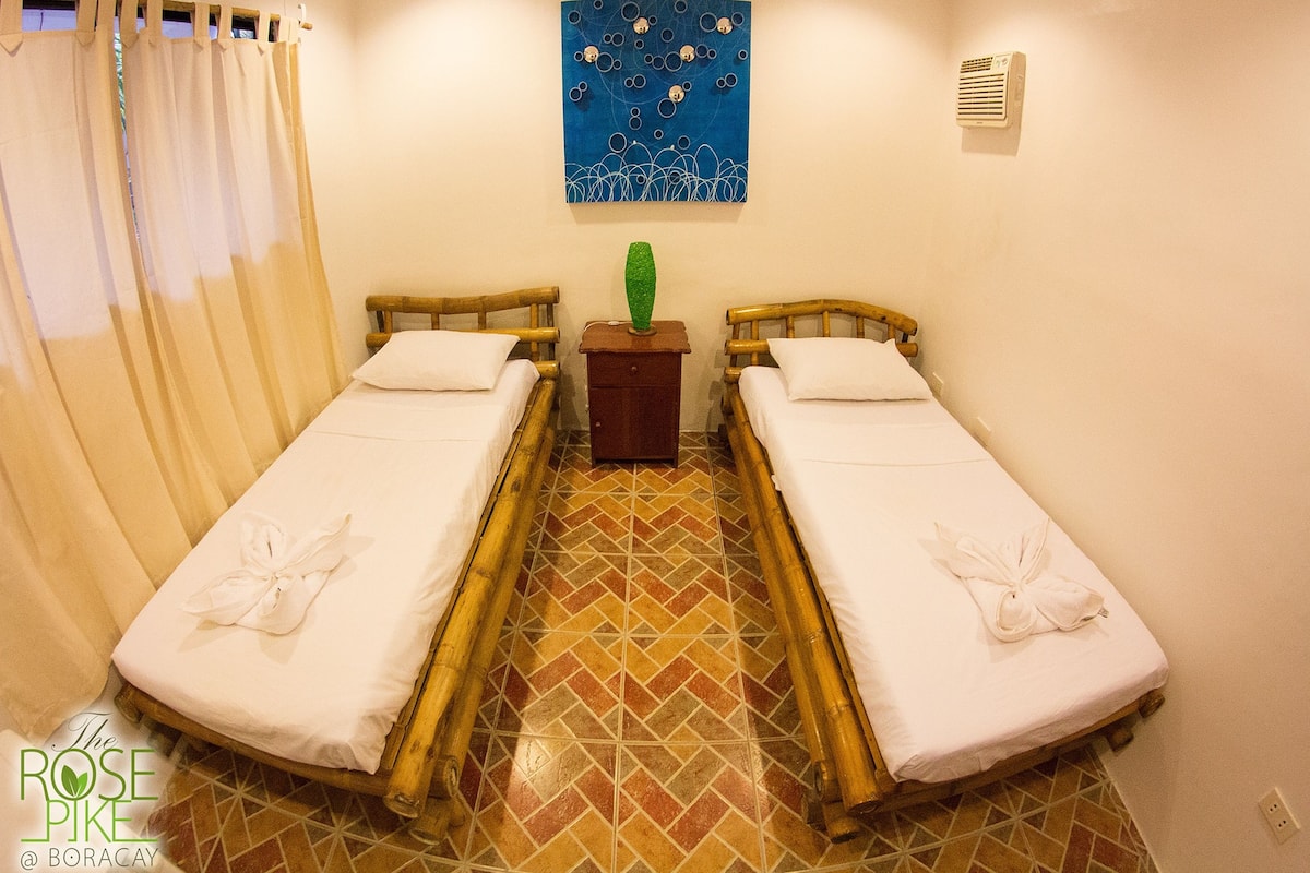 A room with two wooden beds, a small wooden nightstand with a green vase, a blue abstract painting on the wall, and a tiled floor.
