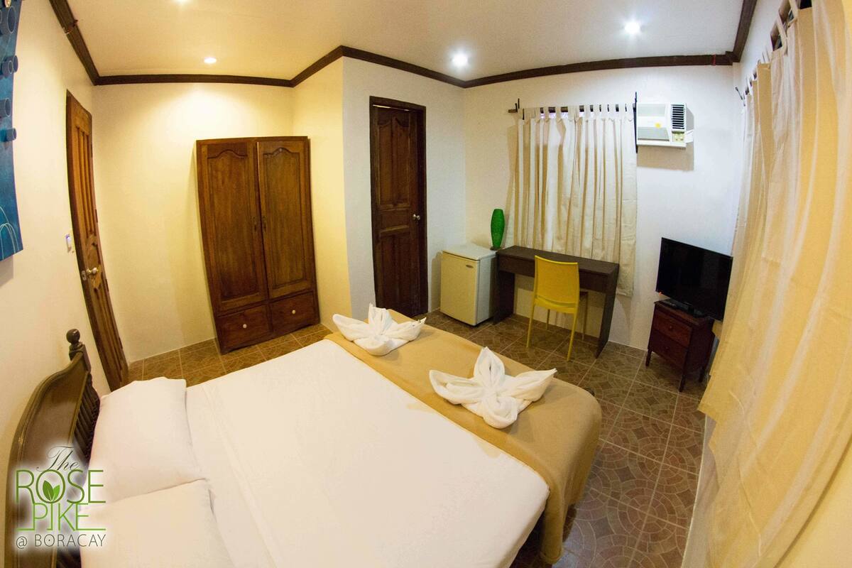 A hotel room with a bed, a desk, a chair, a television, and an air conditioning unit.