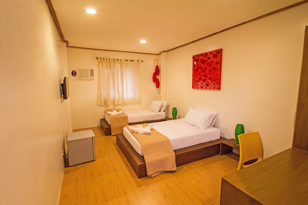 A hotel room with two single beds, a wooden desk, a chair, a small cabinet, and a red abstract painting on the wall.