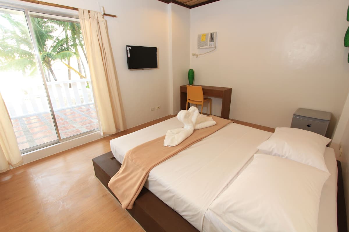 A hotel room with a bed, a desk, a chair, a TV, and a view of palm trees through the window.