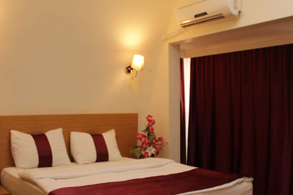 A hotel room with a wooden bed, a red blanket, a wall-mounted air conditioner, and a nightstand with a lamp.
