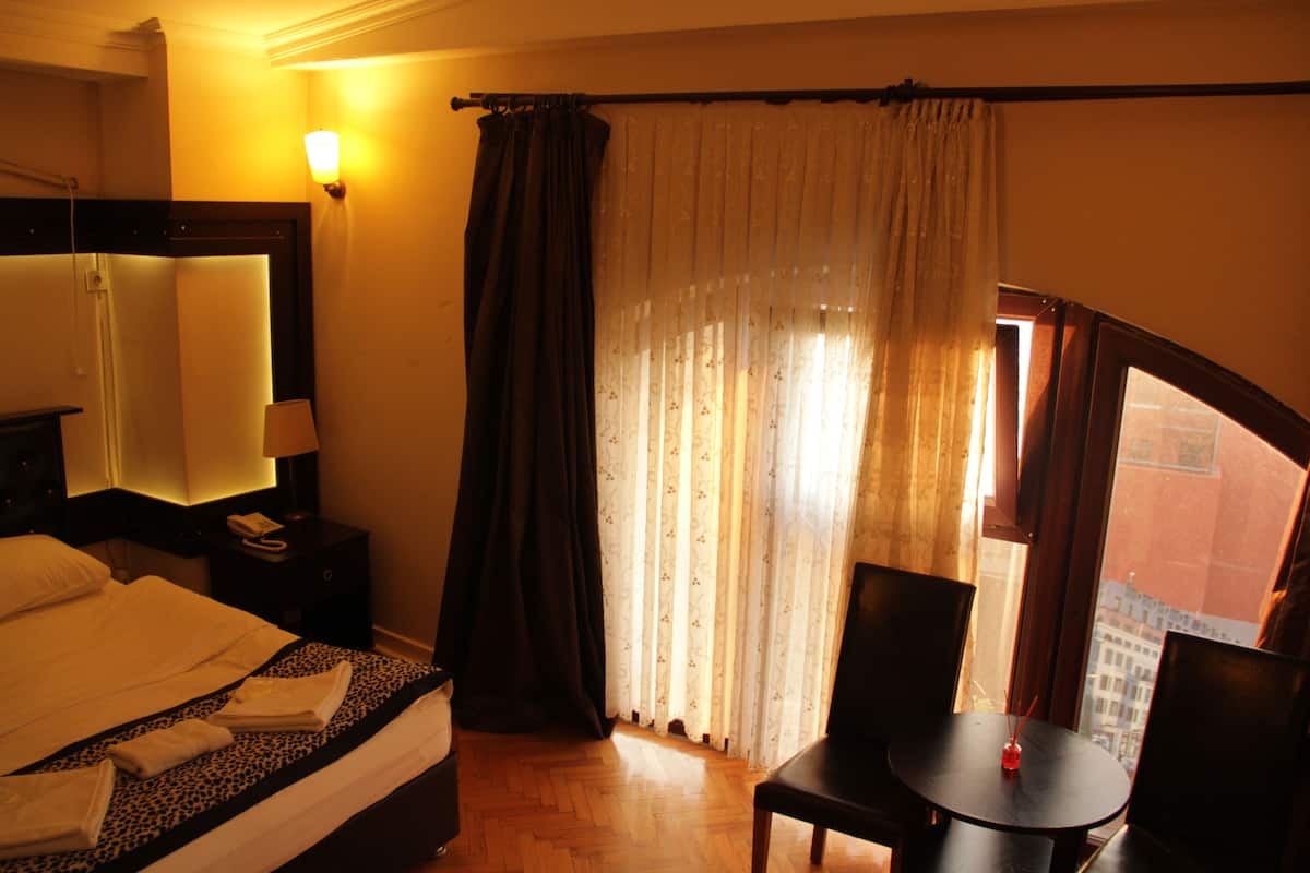 A hotel room with a bed, a table, and chairs, a window with curtains, and a view of buildings outside.