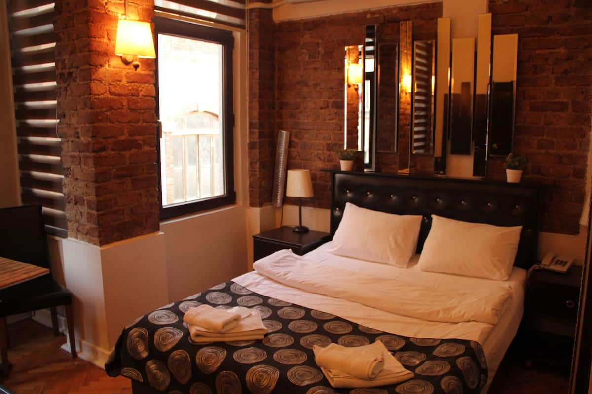 A hotel room with a brick wall, a large bed with a patterned bedspread, a nightstand with a lamp, a mirror, and a window with blinds.