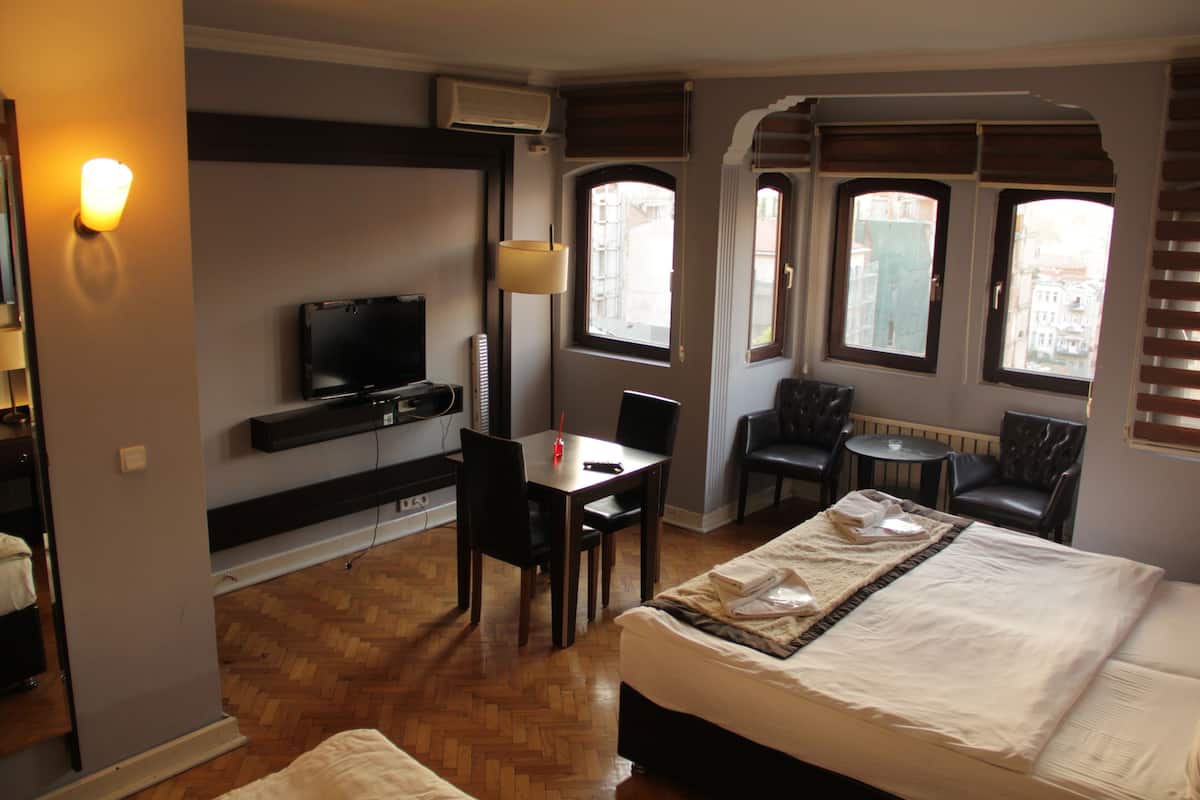 A hotel room with a bed, a desk, a chair, a TV, and windows with views of buildings.
