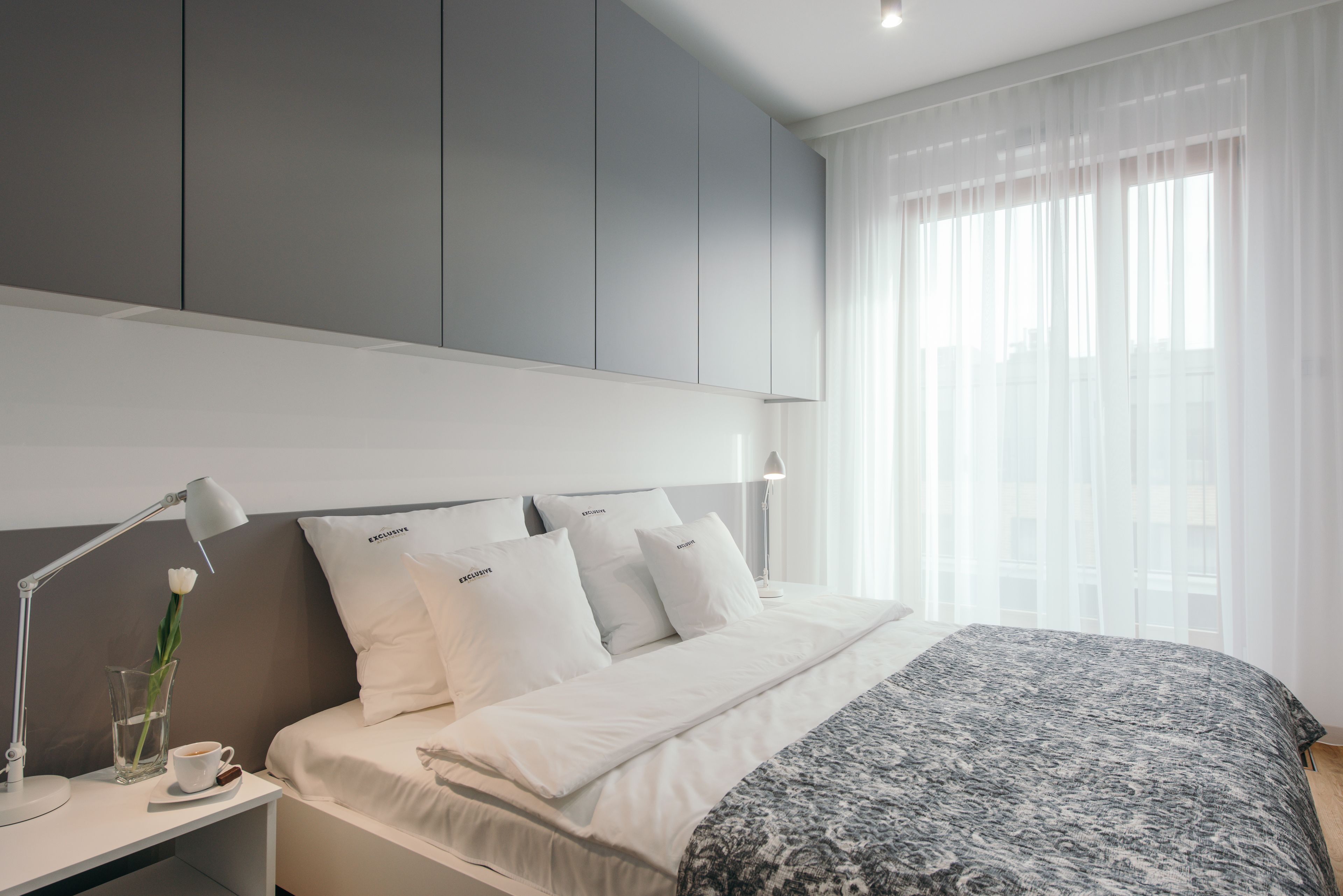 Una habitación de hotel moderna con una cama bien hecha, una mesita de noche con una lámpara y una taza, y un ventanal con cortinas traslúcidas.