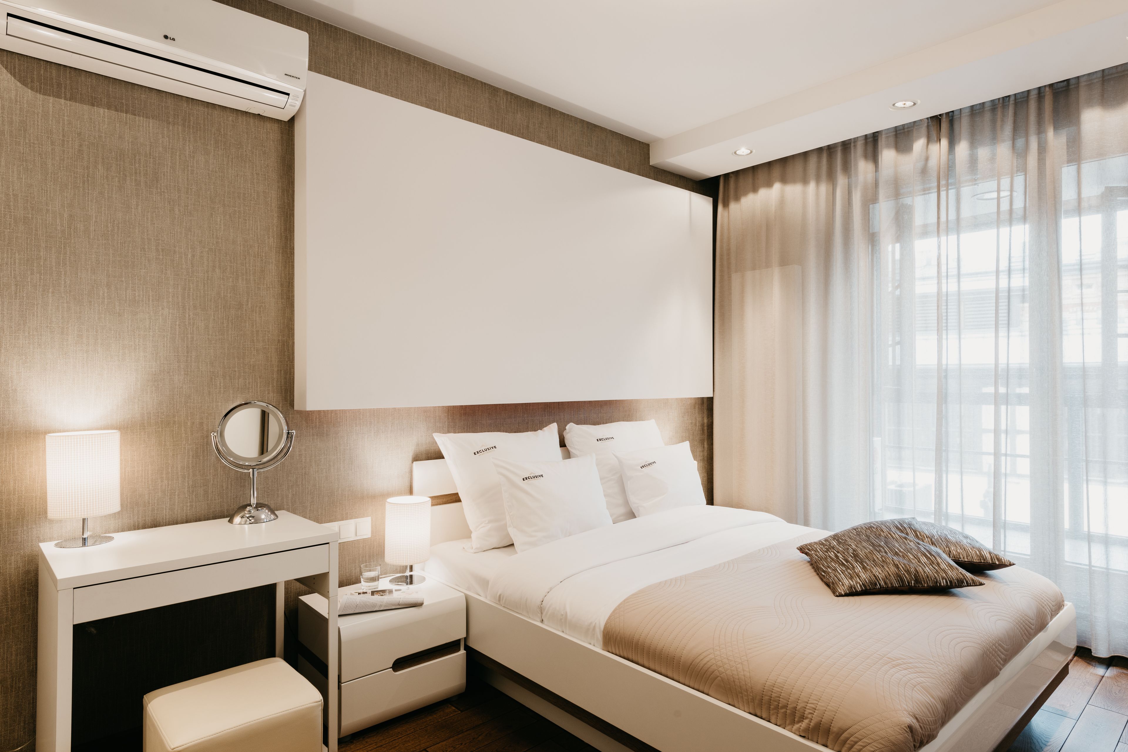 A modern hotel room with a large bed, a bedside table with a lamp, a mirror, and a wall-mounted air conditioner.