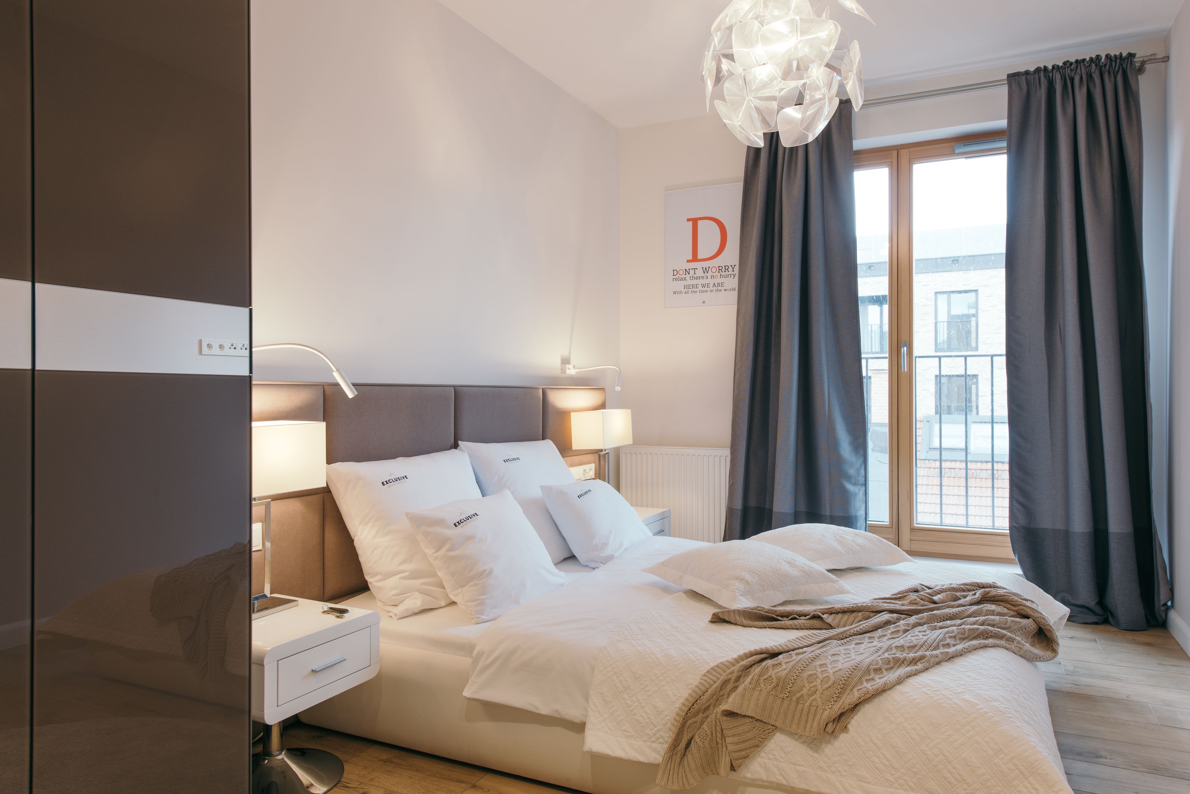 A modern bedroom with a large bed, bedside lamps, a nightstand, and a view of the outside through a window with curtains.