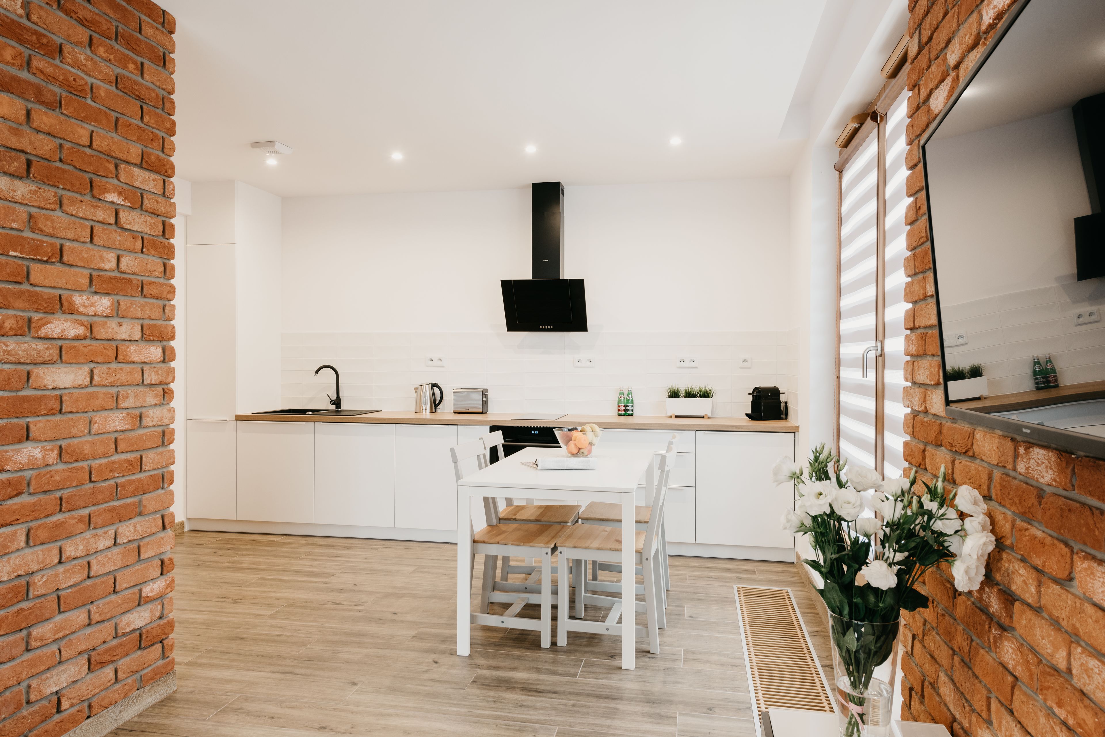 A modern kitchen with white cabinets, a wooden dining table, and a brick accent wall.