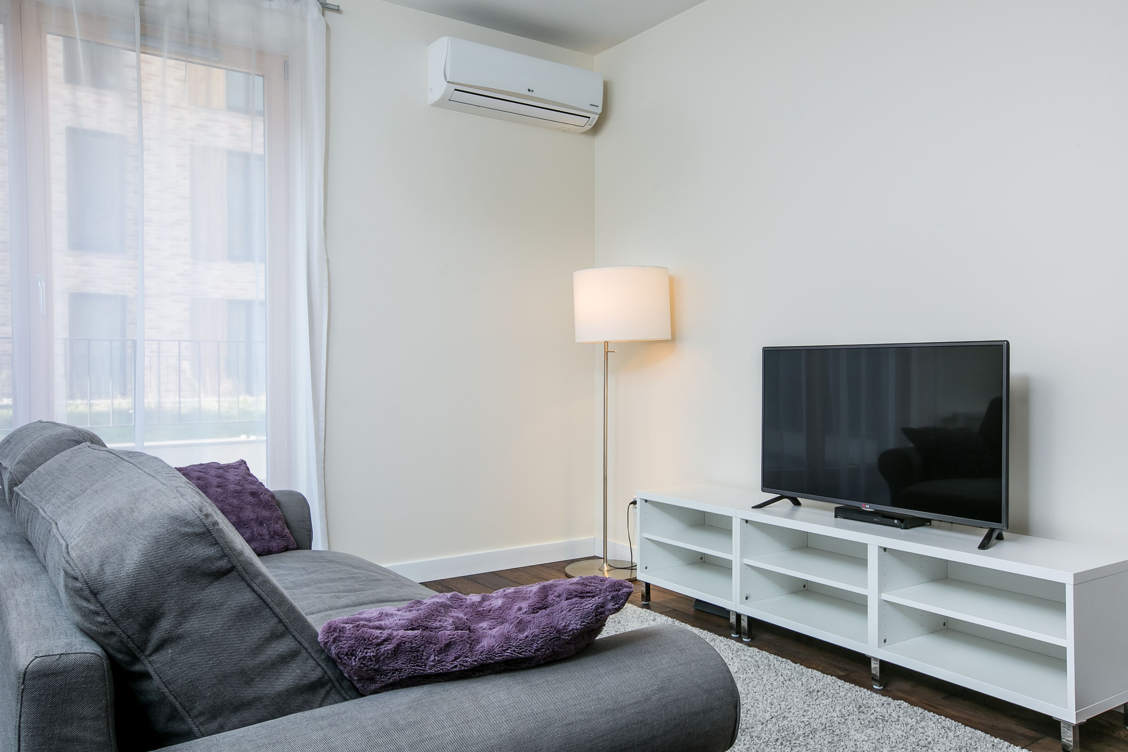 A modern living room with a grey sofa, a flat-screen TV on a white stand, a floor lamp, and an air conditioning unit on the wall.