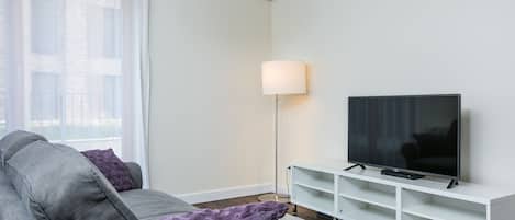A modern living room with a grey sofa, a flat-screen TV on a white stand, a floor lamp, and an air conditioning unit on the wall.
