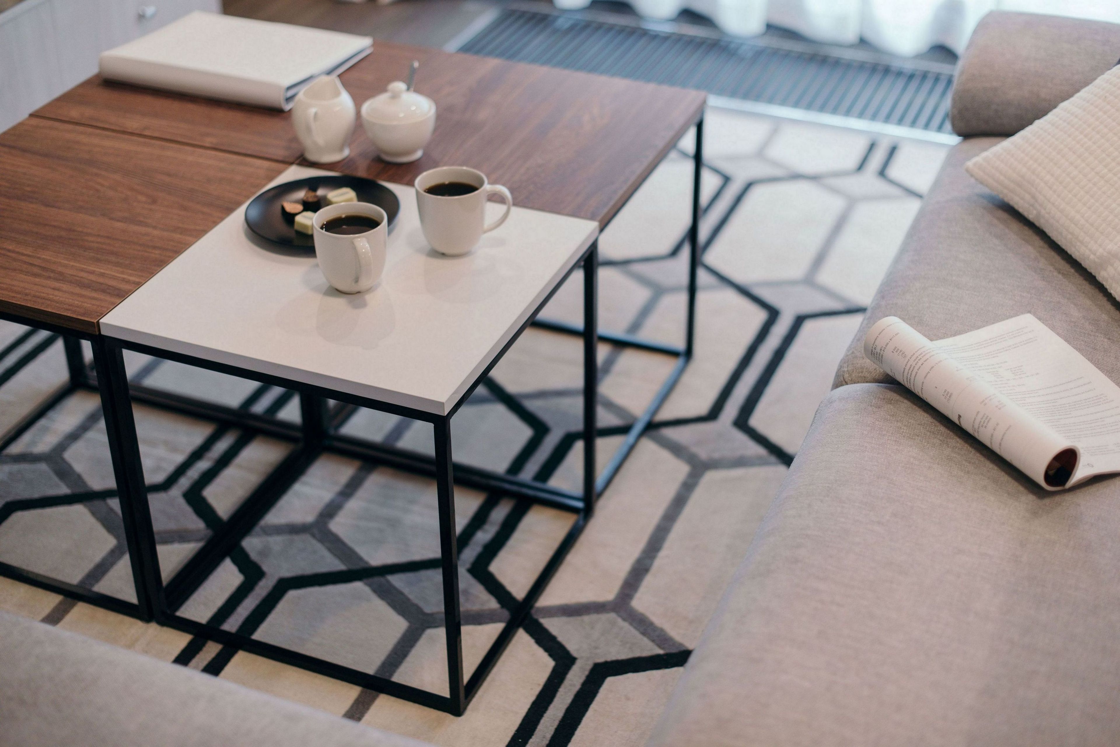 A modern living room with a geometric patterned rug, a wooden coffee table with a white mug and teapot, and a beige sofa with a rolled-up magazine.