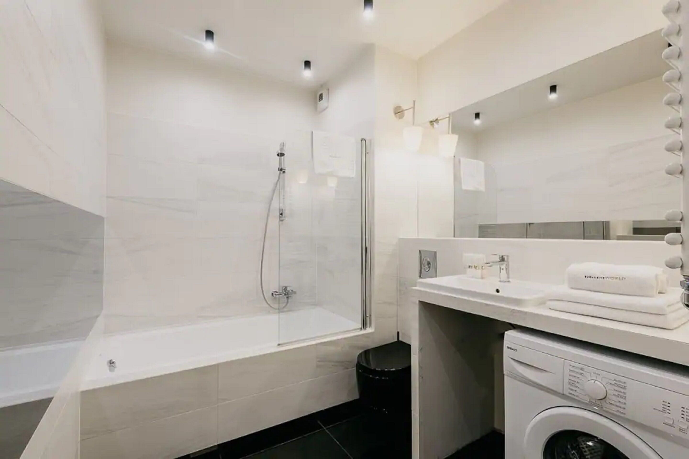 A modern bathroom with a glass-enclosed shower, a white bathtub, a sink with a mirror, and a washing machine.