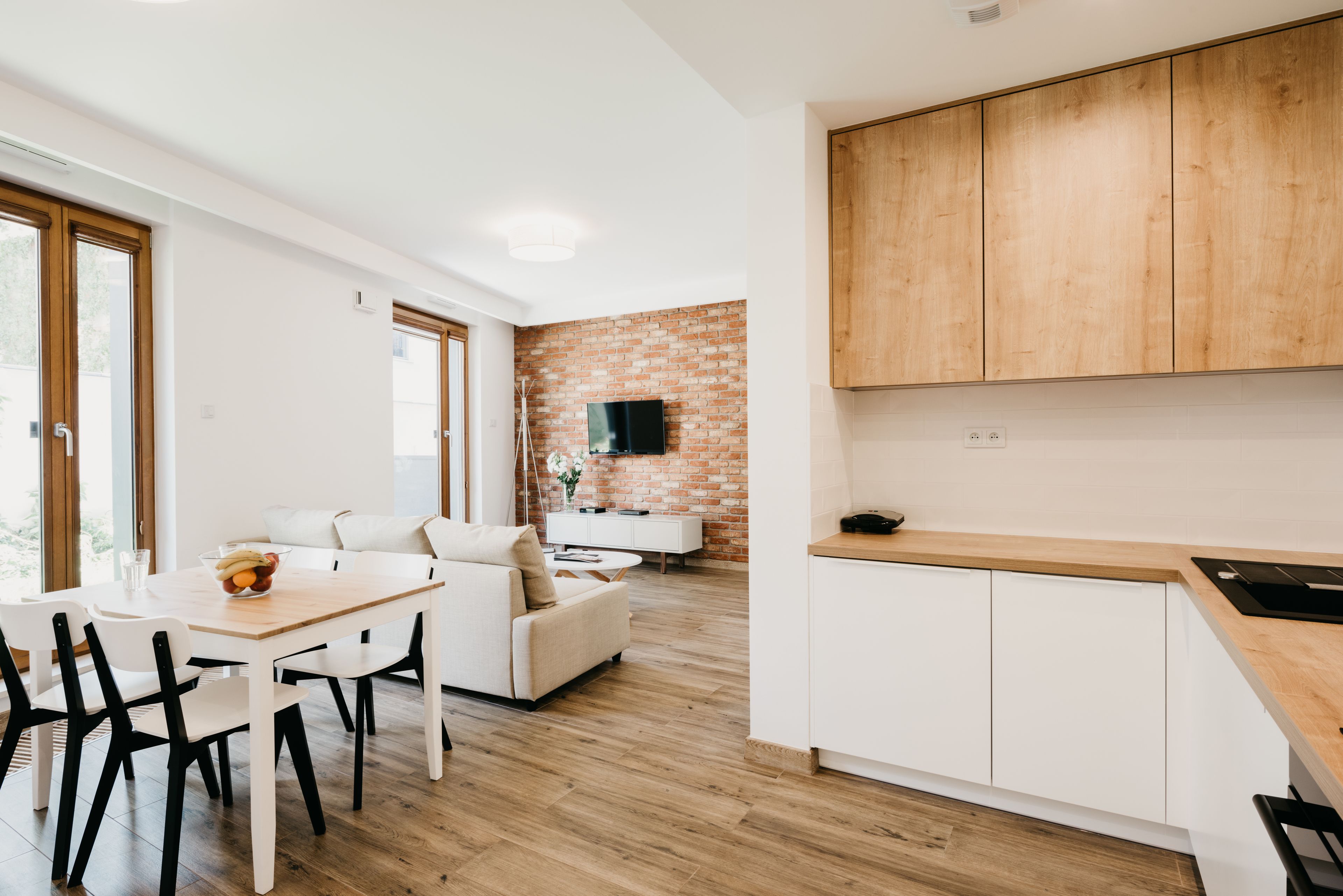 A modern kitchen with wooden cabinets, a dining area with a white table and chairs, and a brick accent wall.