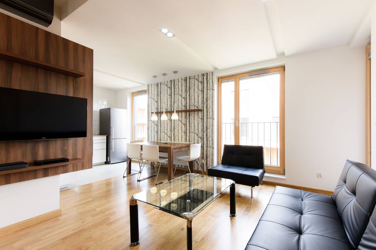 A modern living room with a glass coffee table, a black sofa, a dining area with a wooden table and chairs, and a TV mounted on a wooden panel.