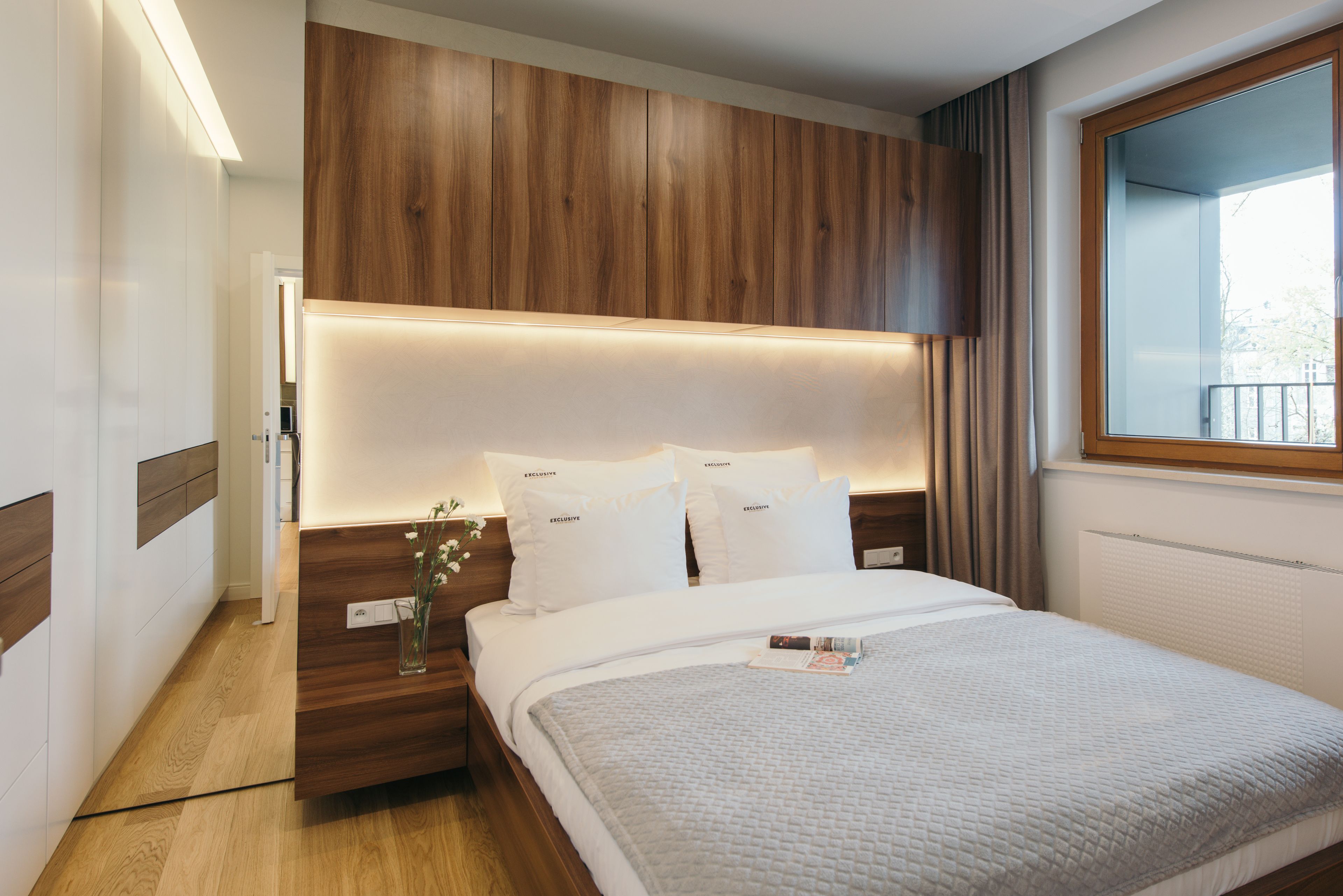 A modern hotel room with a wooden headboard, a bedside table with a vase of flowers, and a large window with curtains.