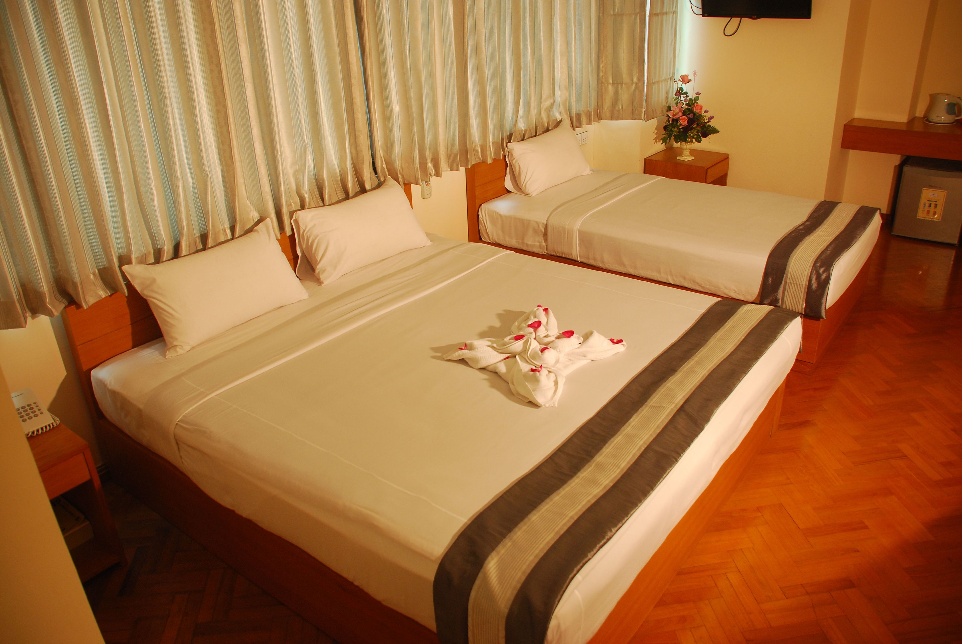 A hotel room with two beds, a wooden floor, a TV on the wall, and a small table with a vase of flowers.