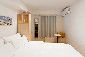 A hotel room with a bed, a small table, a seating area with cushions, a wardrobe, a mirror, and an air conditioning unit. - Hotel Marguerite (Seoul)