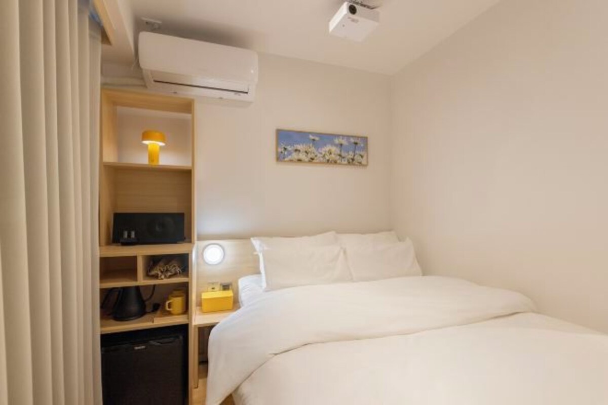 Habitació d'hotel amb un llit, una taula de nit amb una làmpada, una estant amb una làmpada, un escriptori petit amb una làmpada i un quadre a la paret.