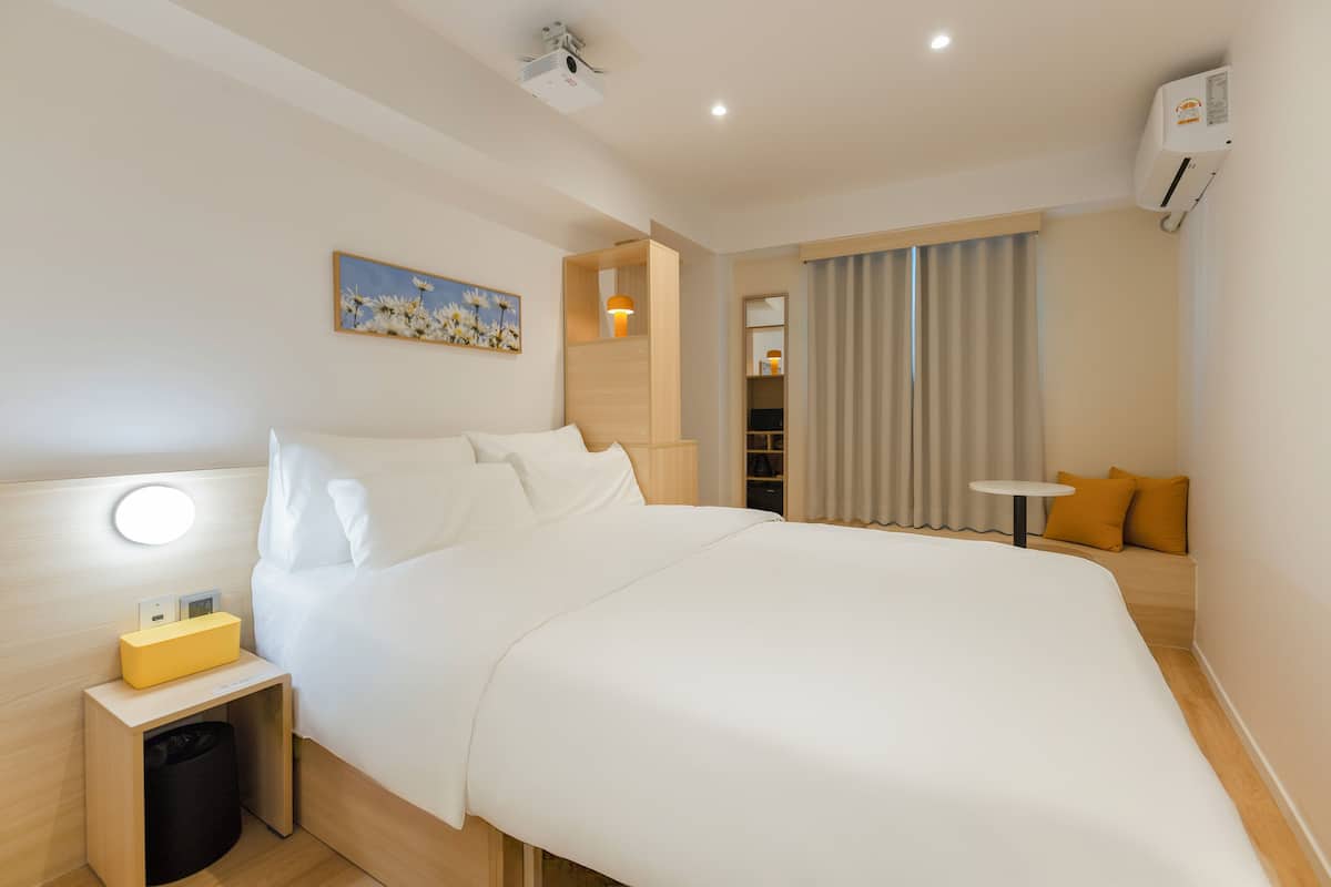 Une chambre d’hôtel avec un grand lit, une table de chevet avec une lampe, une petite table, une poubelle, une armoire et une fenêtre avec des rideaux.