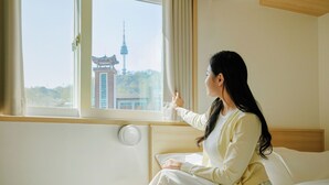 Une personne est assise près d’une fenêtre, dans une pièce qui contient un lit, une table de chevet et un thermostat fixé au mur.