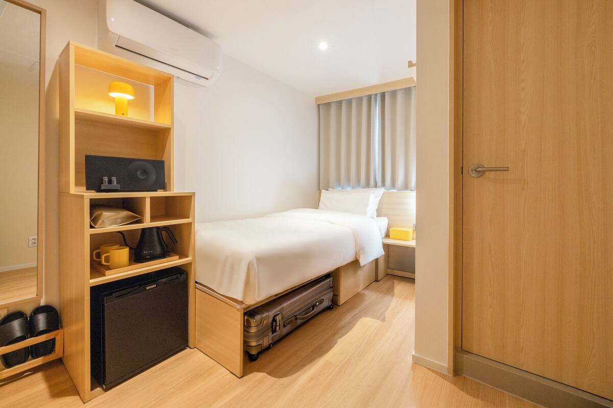 Une chambre d’hôtel compacte comprenant un lit, une petite kitchenette, un espace de rangement intégré et une fenêtre avec des rideaux.