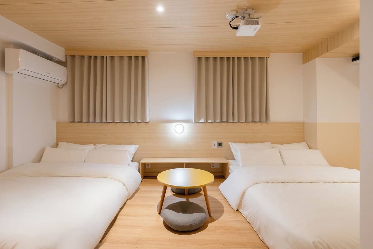 Une chambre d’hôtel avec deux lits, une petite table ronde, un banc et un projecteur au plafond.