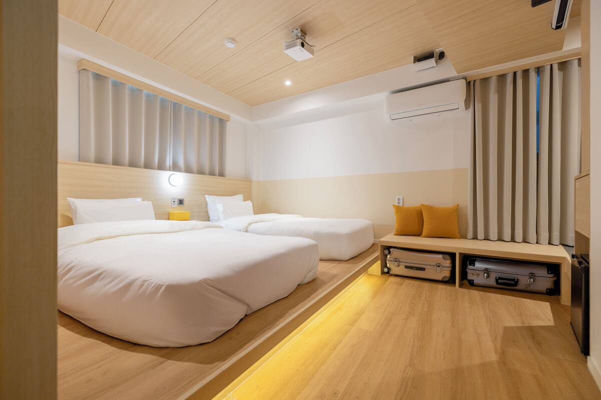 Une chambre d’hôtel avec un plafond en bois, deux lits, un banc avec espace de rangement et un appareil de climatisation.