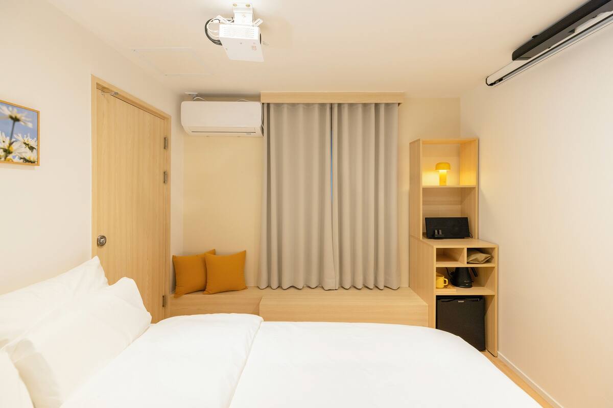 Une chambre d’hôtel comprenant un lit, un bureau avec une lampe, un appareil de climatisation et une étagère avec divers objets.