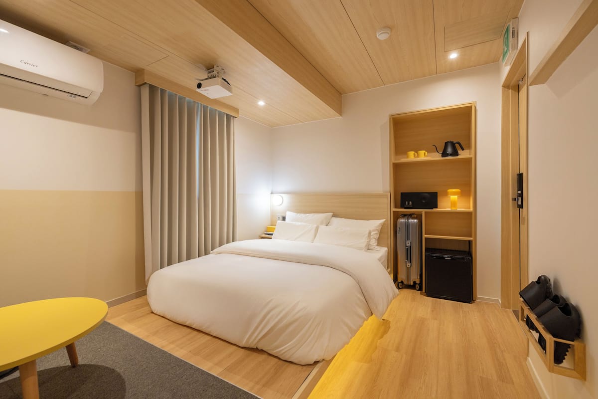 Une chambre d’hôtel comprenant un lit, une petite table, un bureau, une armoire et une étagère avec des articles de toilette.
