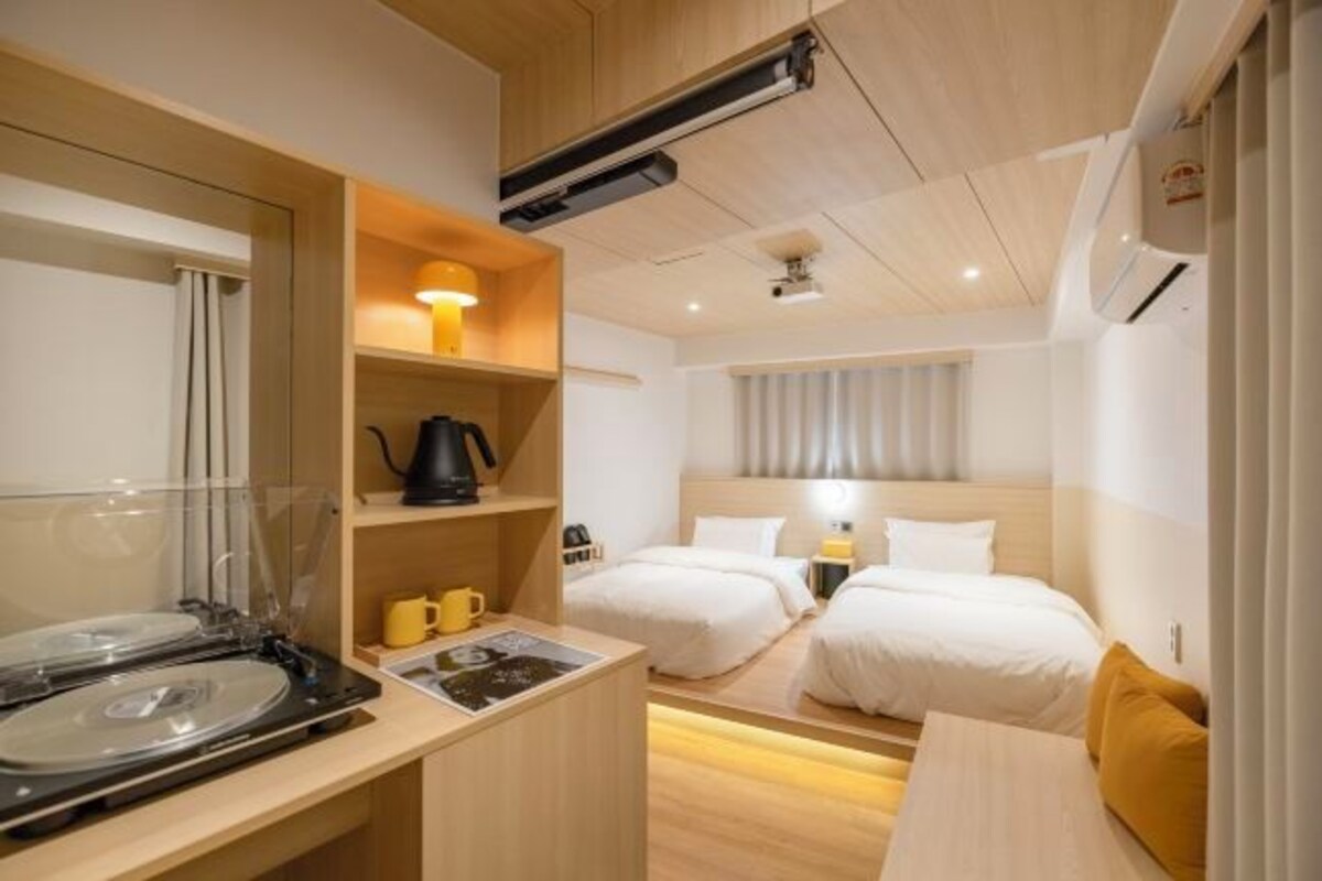 Habitació d'hotel senzilla amb dos llits individuals, una cuineta petita, zona de menjador i aire condicionat fixat al sostre.