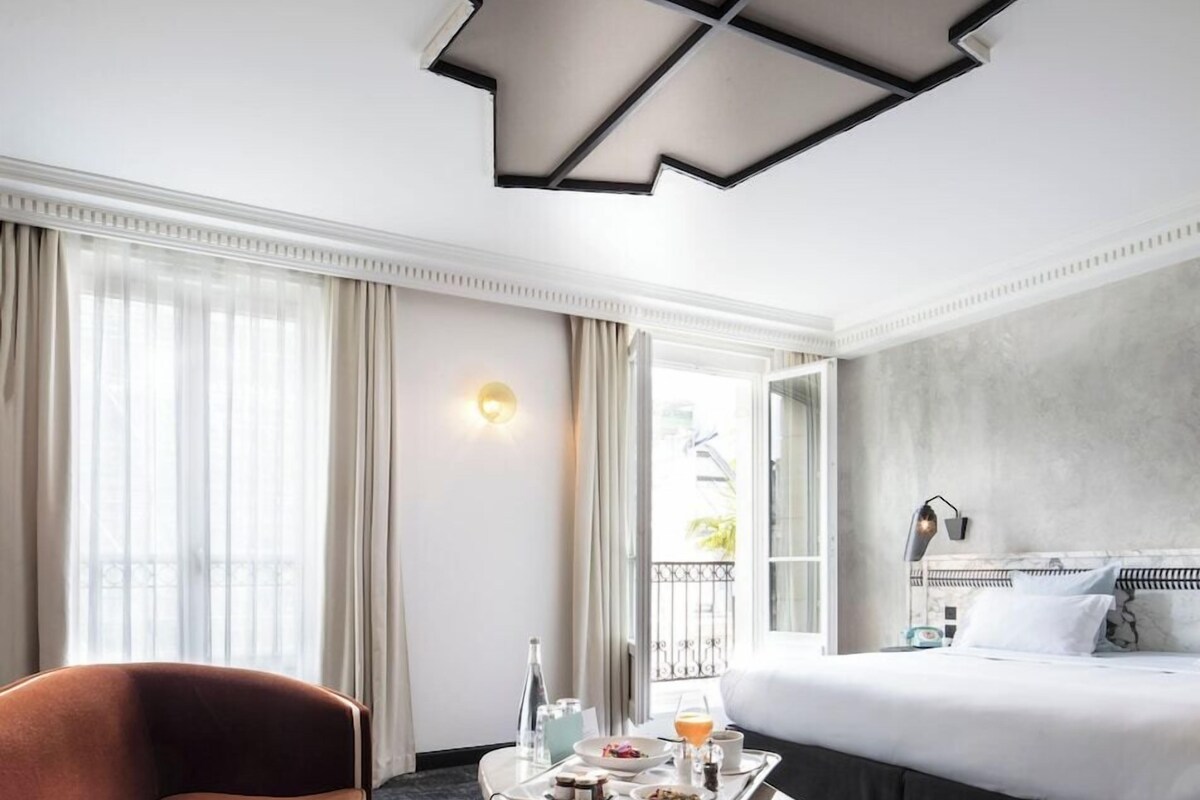 A hotel room with a bed, a chair, a table with breakfast items, a lamp, and a view of the outside.